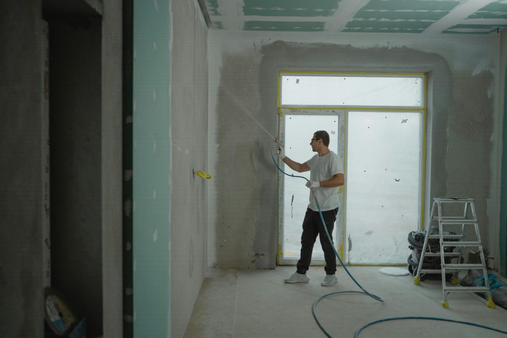 pexels photo 6474133 6474133 A man sprays paint on a wall while renovating an indoor room with professional equipment.